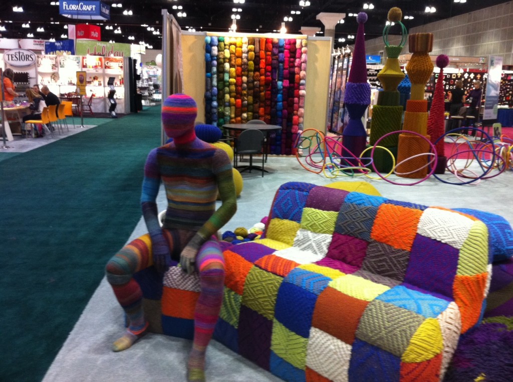Yarn man, sitting on a patchwork with additional yarn creations in the background